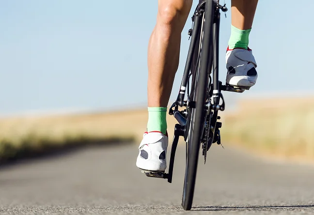 Les jambes d'un cycliste, chaussées de chaussures blanches et de chaussettes vert fluo, pédalent sur un vélo noir sur une route dégagée sous un ciel bleu limpide, évoquant le mouvement et la liberté en plein air
