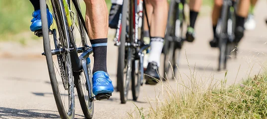 Des cyclistes sur une piste ensoleillée, l'accent étant mis sur leurs jambes et leurs chaussures de cyclisme bleues. La scène dégage une impression de mouvement et d'énergie, avec de l'herbe qui borde la route