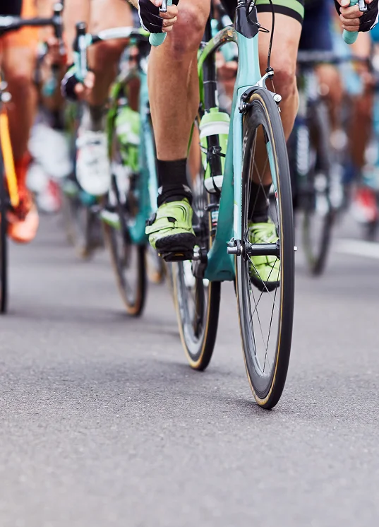 Des cyclistes participant à une course sur route, dont l'accent est mis sur leurs jambes et leurs vélos colorés, roulent en peloton serré dans une rue. La scène dégage une impression de vitesse, de compétition et de travail d'équipe