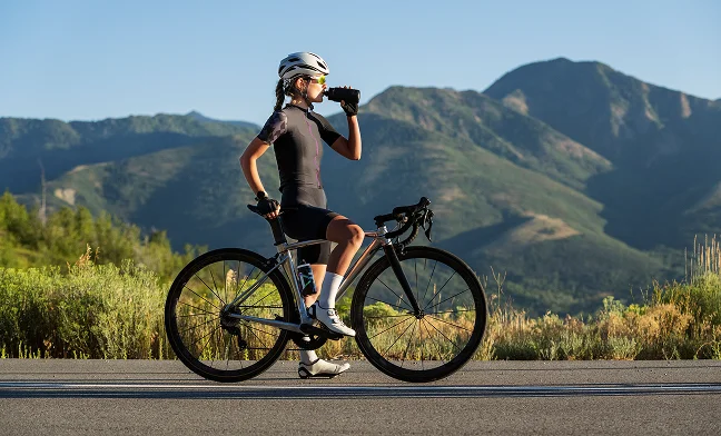 Cycliste sportive portant un casque et une tenue de vélo, arrêtée sur une route de montagne et buvant dans une gourde, avec des montagnes verdoyantes en arrière-plan