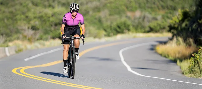 Un cycliste vêtu d'un maillot violet gravit une route sinueuse et ensoleillée, bordée de collines verdoyantes. La scène dégage une impression de détermination et de concentration